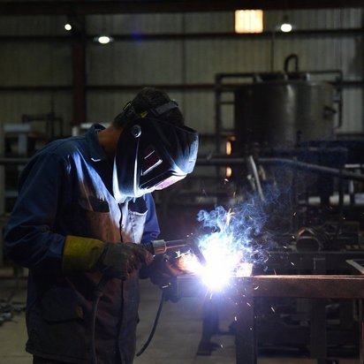 Vraag een offerte aan bij TÜV NORD voor ISO 3834 certificering Lasser met beschermende helm aan het lassen van een metalen constructie in een werkplaats.