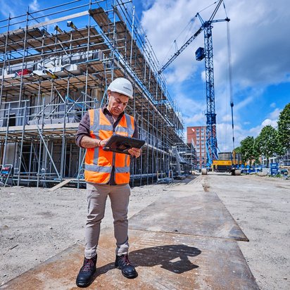 Vraag een offerte aan bij TÜV NORD voor VCU certificering Bouwinspecteur met helm en tablet controleert bouwplaats met steigers en hijskraan.